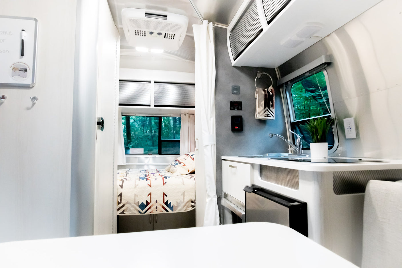 Cozy, modern RV rental interior with a compact kitchen, comfortable bedroom featuring a patterned quilt, and scenic forest views.