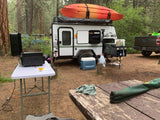 Compact RV rental set up at a forest campsite, featuring a travel trailer with a kayak, grill, and truck.