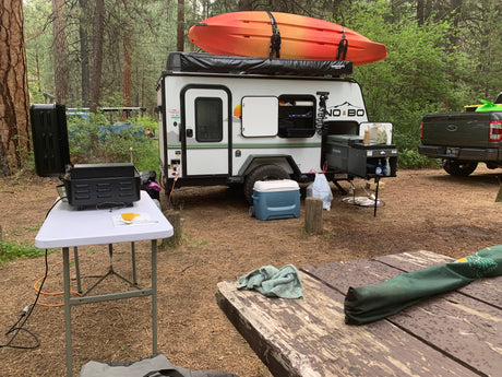 Compact RV rental set up at a forest campsite, featuring a travel trailer with a kayak, grill, and truck.