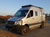 Custom silver Mercedes Sprinter RV rental parked outdoors. This rugged adventure camper van features off-road tires, a winch, and roof rack.