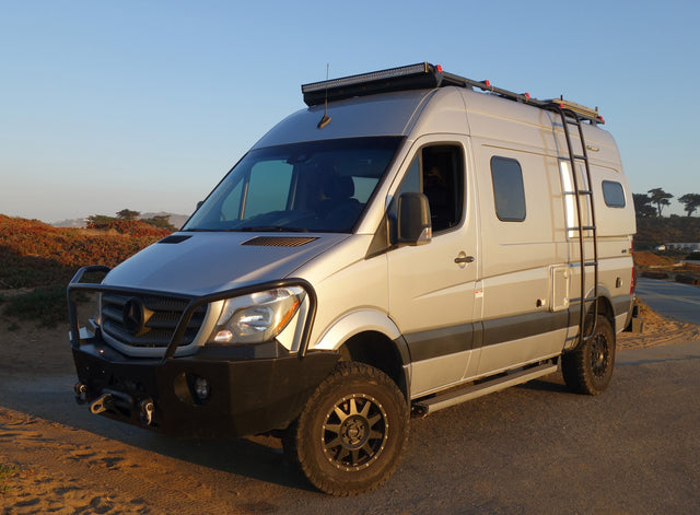 Custom silver Mercedes Sprinter RV rental parked outdoors. This rugged adventure camper van features off-road tires, a winch, and roof rack.