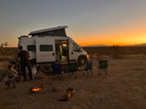 A white RV rental camper van in the desert at sunset. A family cooks by a fire, chairs set up for a relaxing remote camp.