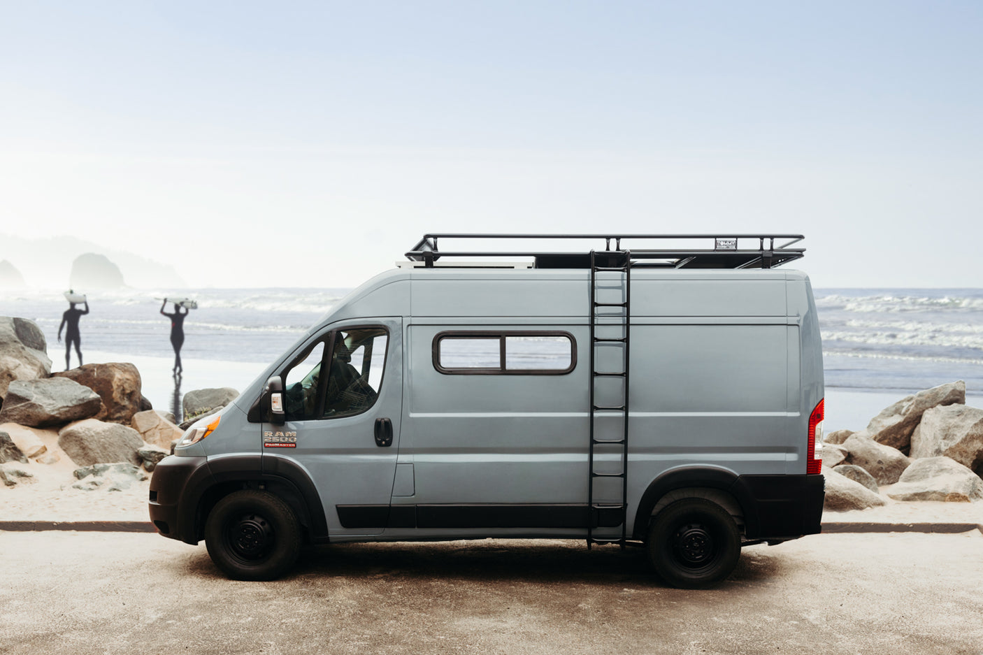 This grey RV rental van, equipped with a roof rack and ladder, is perfectly set on a beach with surfers nearby.