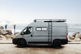 This grey RV rental van, equipped with a roof rack and ladder, is perfectly set on a beach with surfers nearby.