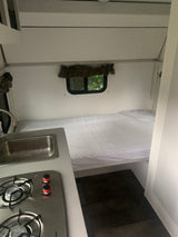 Compact RV rental interior featuring a comfy bed, window with nature views, and a small kitchen with sink and stovetop.