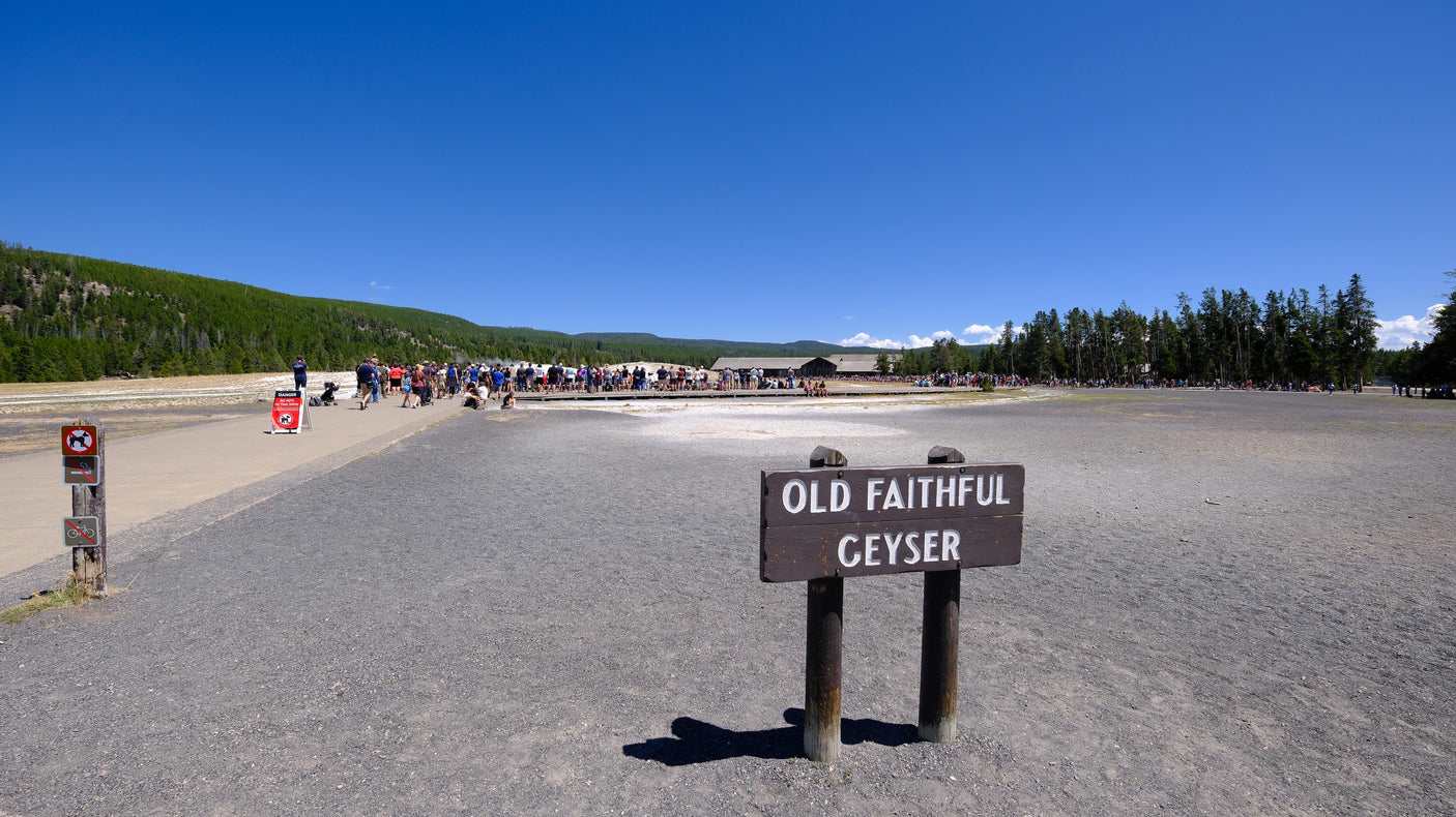 The "OLD FAITHFUL GEYSER" sign stands before a large crowd gathered in Yellowstone National Park. Plan your rv rental trip!