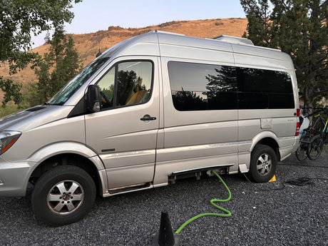 Silver Roadtrek RV rental (camper van) at a gravel campsite with hookups, bikes, and mountain views.