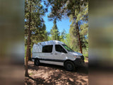White RV rental van parked among tall pines under a blue sky, perfect for a forest escape.