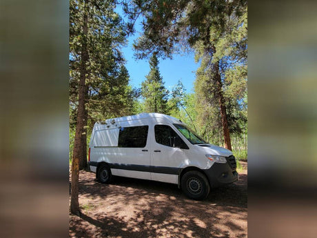 White RV rental van parked among tall pines under a blue sky, perfect for a forest escape.