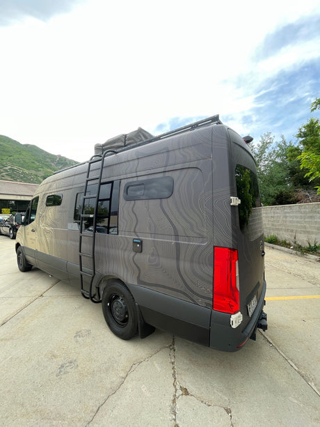 Adventure-ready gray Sprinter RV rental with topographic map wrap, roof rack, & side ladder for off-grid travel.