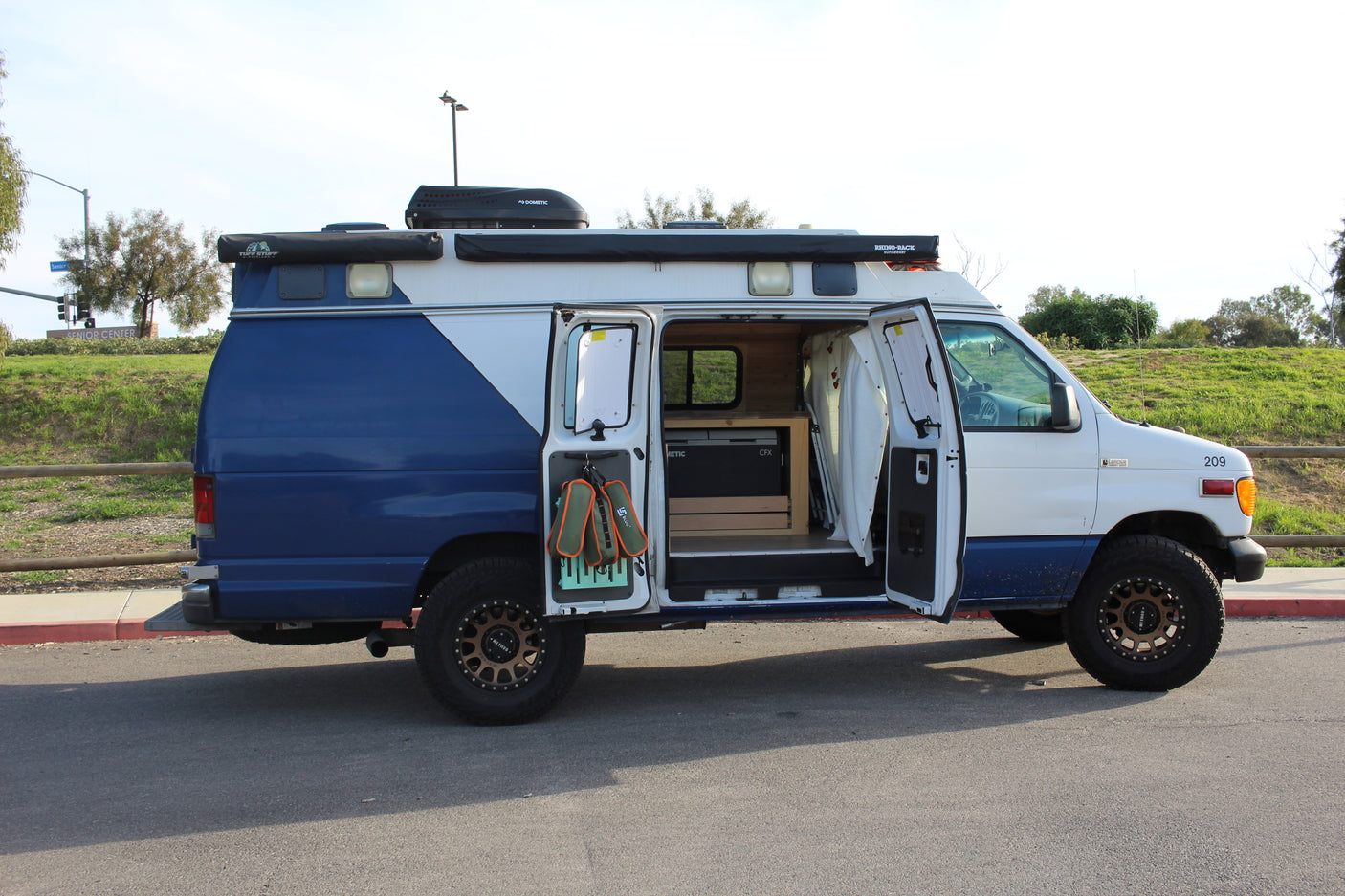 Side view of a blue/white Ford camper van. Interior and roof gear visible. Perfect for an RV rental adventure.