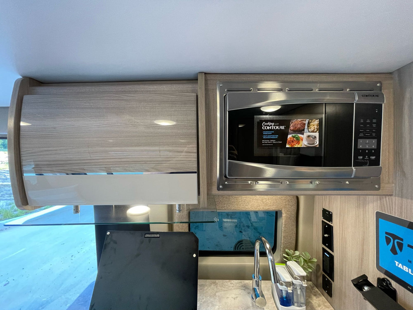 This RV rental kitchen features a sleek Contoure microwave, sink, modern cabinetry, and a tablet display. — photo 12