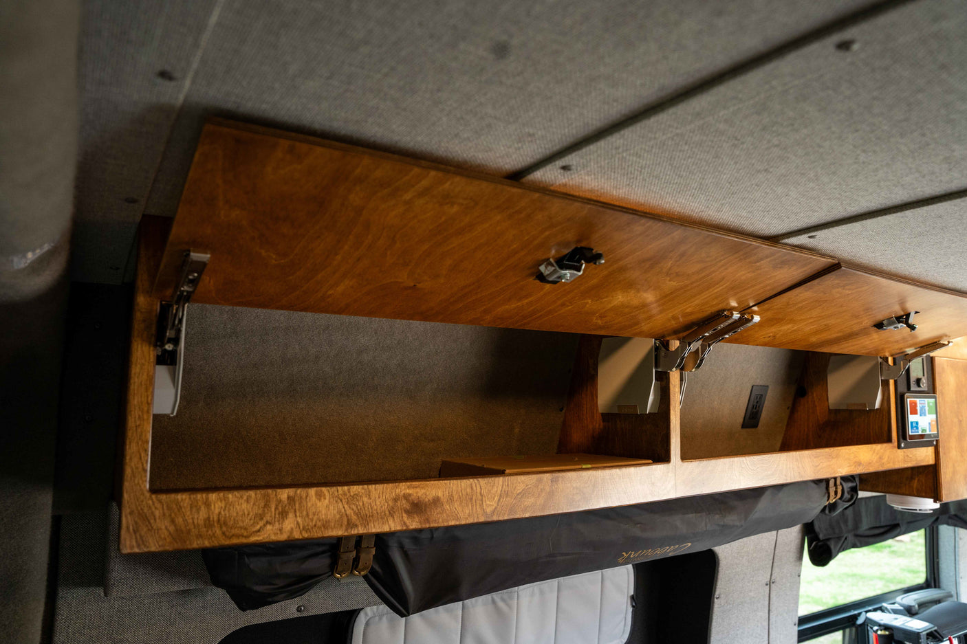 RV rental interior: Open wooden overhead cabinets with gas struts, showing storage, a control panel, and fabric-lined walls.
