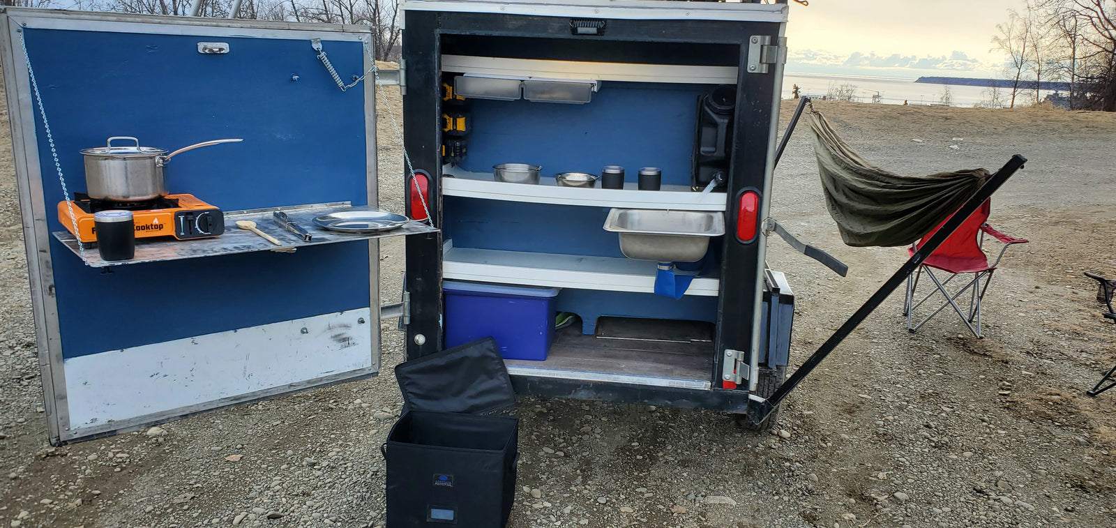 Compact RV rental with an innovative outdoor kitchen featuring a portable stove, pot, sink, and shelves for roadside cooking by the water.