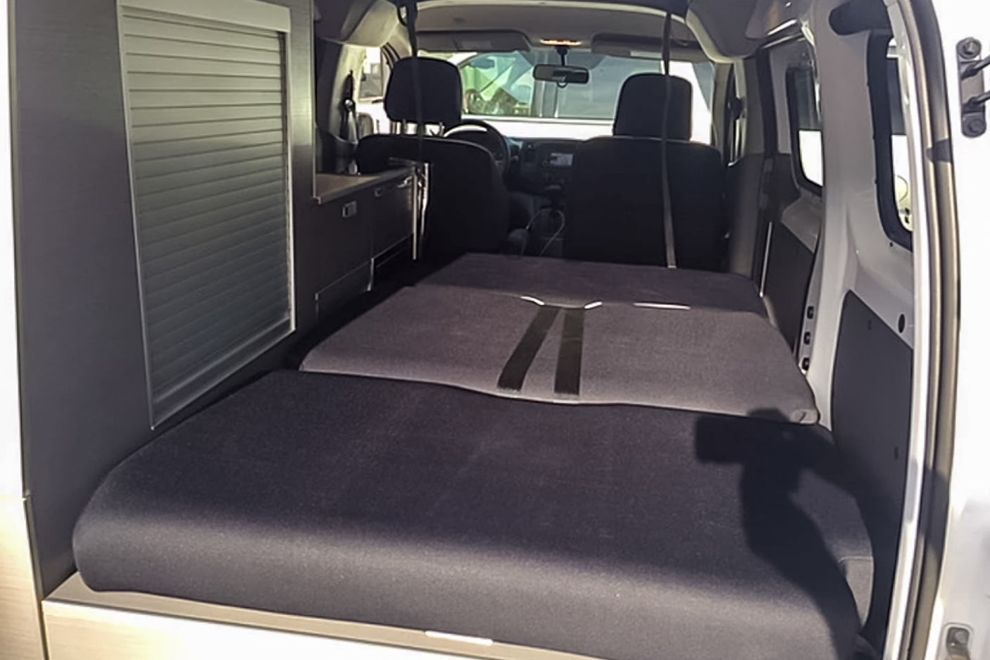Inside an RV rental: a flexible dark grey bed system unfolds towards the front seats, with sleek storage cabinets on the left.