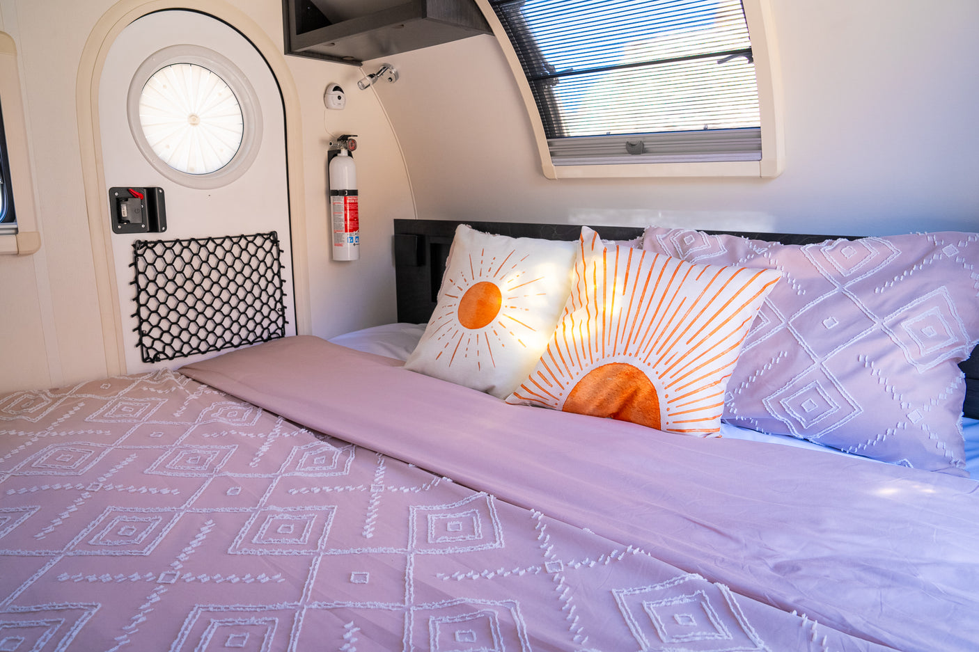 RV rental bedroom featuring a comfy bed with pink bedding, vibrant sun pillows, a round door, and a safety extinguisher.