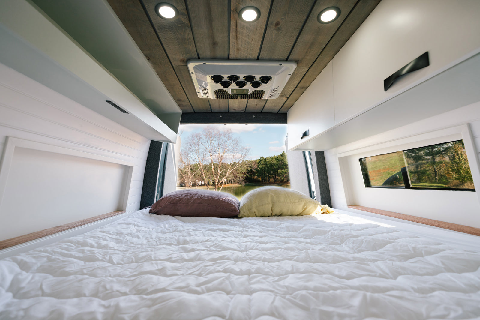 Cozy RV rental interior: white bed, pillows, modern cabinets, and a scenic lake view through the rear window.