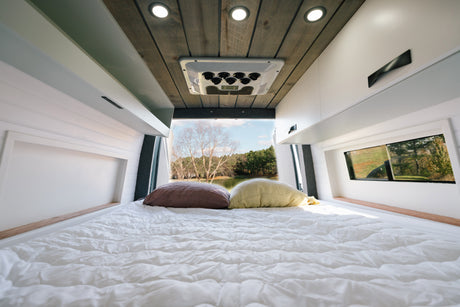 Cozy RV rental interior: white bed, pillows, modern cabinets, and a scenic lake view through the rear window.