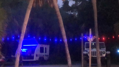 RV rental illuminated by blue interior lights, parked at campsite 28 with a Jeep, framed by palms & festive string lights.
