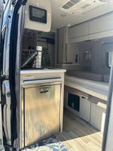 Inside a modern rv rental: stainless fridge, counter with supplies, and comfortable bed/seating. Clean, well-lit interior.