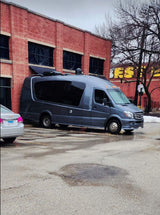A sleek gray RV rental, a Mercedes Sprinter conversion van, is parked by a red brick building in a wet lot.
