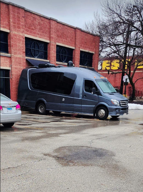 A sleek gray RV rental, a Mercedes Sprinter conversion van, is parked by a red brick building in a wet lot.