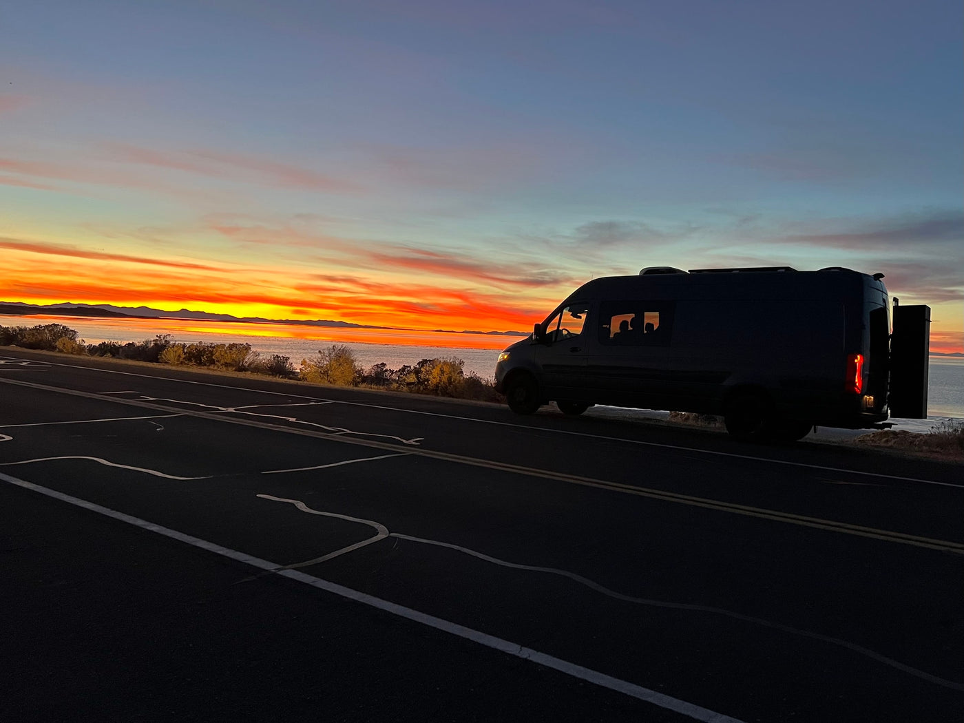 A dark RV rental camper van rests on a scenic roadside, silhouetted against a breathtaking orange sunset over a lake and distant mountains. — photo 2