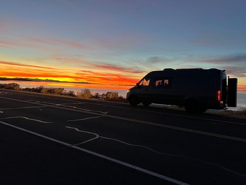 A dark RV rental camper van rests on a scenic roadside, silhouetted against a breathtaking orange sunset over a lake and distant mountains. — photo 2