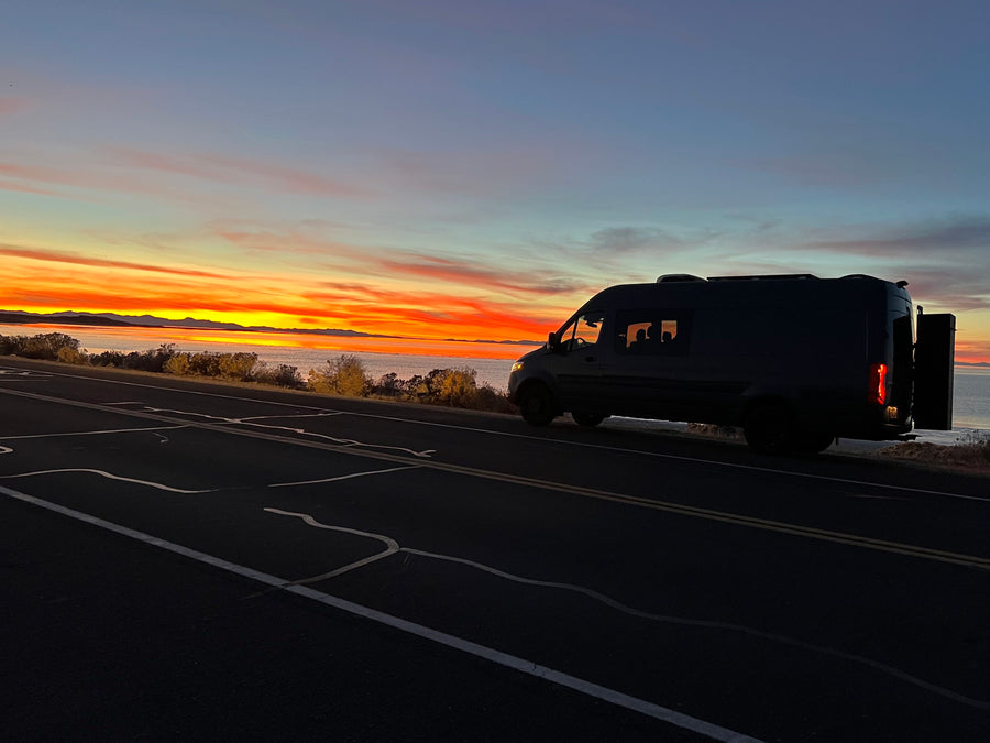 A dark RV rental camper van rests on a scenic roadside, silhouetted against a breathtaking orange sunset over a lake and distant mountains.