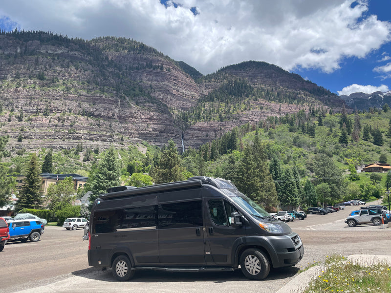 This dark gray RV rental camper van is parked, facing a majestic rocky mountain with trees and a visible waterfall under a cloudy sky. — photo 3
