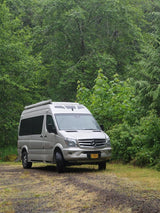 Silver Mercedes Sprinter camper van, an ideal RV rental, parked on a dirt road amidst lush green forest.
