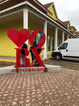 Two people pose with the red LOVE BX sign (@SUPERWINGSOBX) outside a yellow building, a commercial van parked for an RV rental trip start.