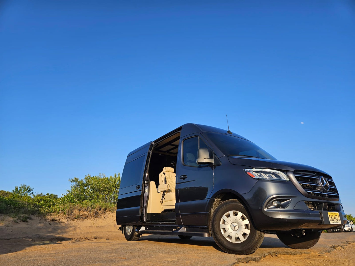Luxury Mercedes Sprinter van, perfect for an RV rental. Parked on a dirt road, side door open, revealing a comfy interior.