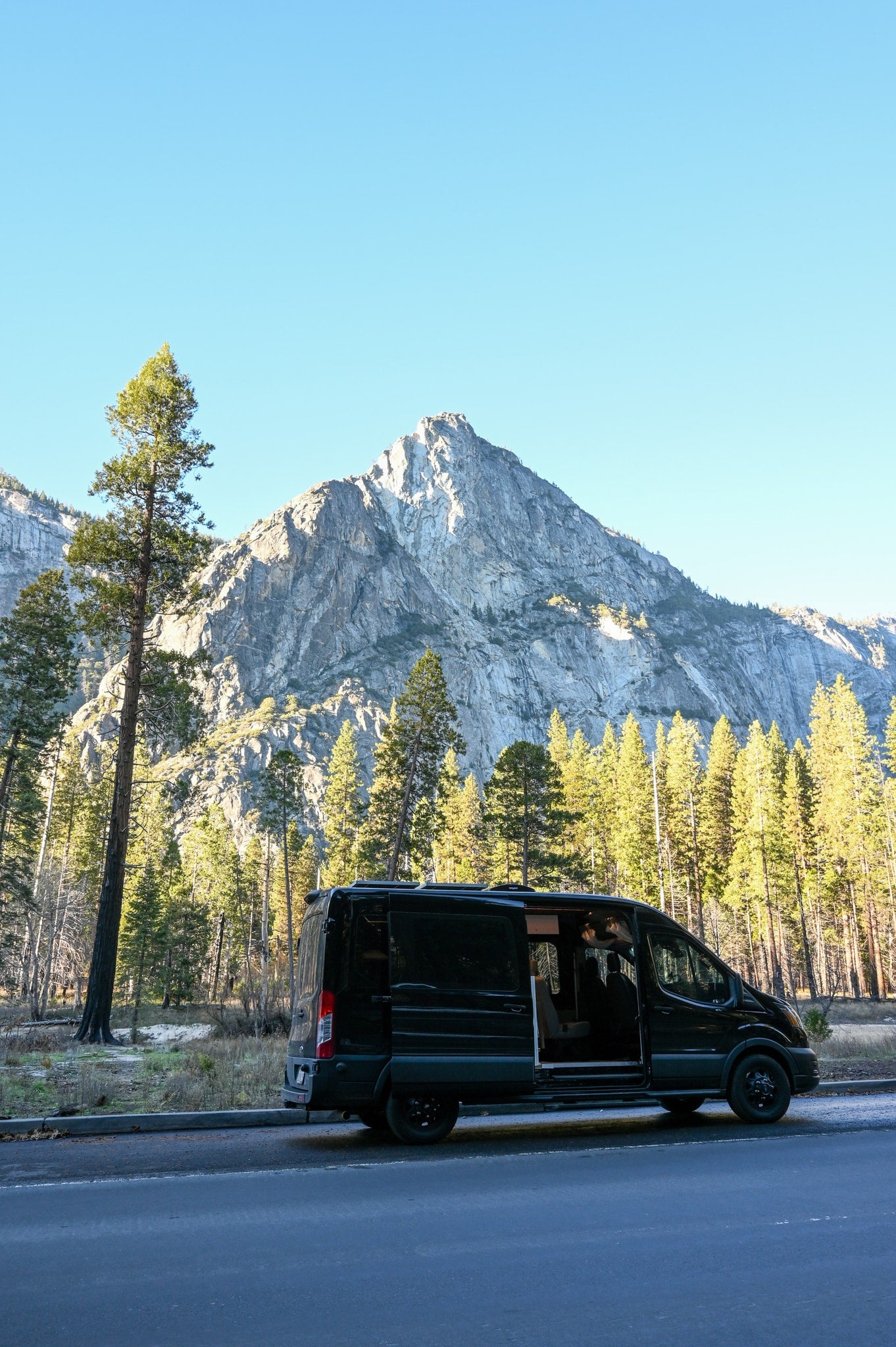A black RV rental van with an open side door is parked on a roadside, against a backdrop of towering pine trees and a grand mountain. — photo 3