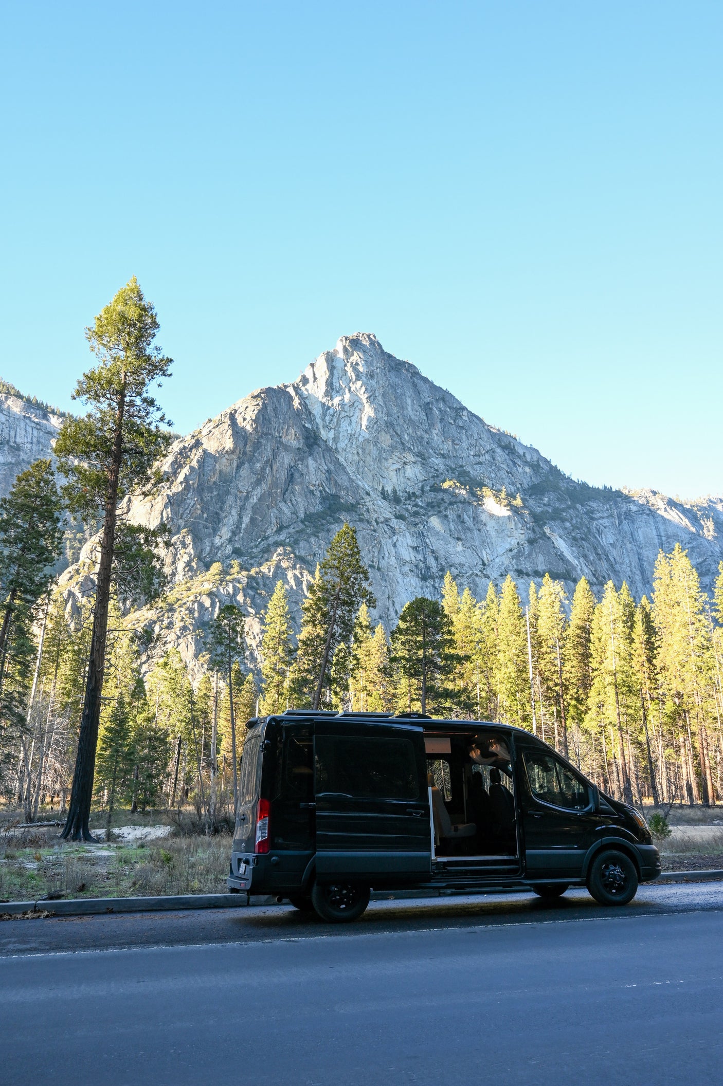 A black RV rental van with an open side door is parked on a roadside, against a backdrop of towering pine trees and a grand mountain.