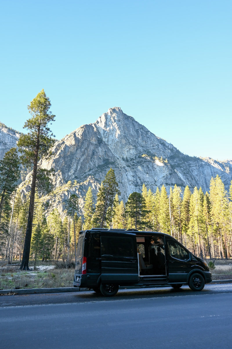A black RV rental van with an open side door is parked on a roadside, against a backdrop of towering pine trees and a grand mountain. — photo 3