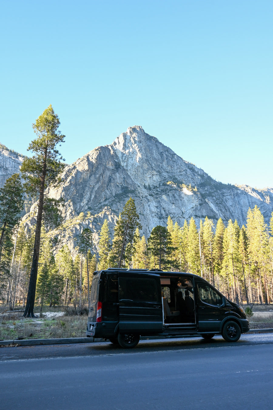 A black RV rental van with an open side door is parked on a roadside, against a backdrop of towering pine trees and a grand mountain.