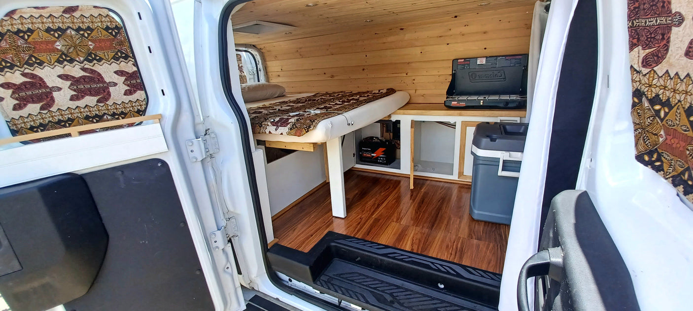 Cozy RV rental interior: wood-paneled camper van with a bed, stove, and cooler, seen through open side door.