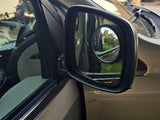 An RV rental's side mirror with a convex blind spot mirror, reflecting outdoor scenery and showing vehicle interior.