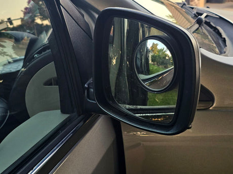 An RV rental's side mirror with a convex blind spot mirror, reflecting outdoor scenery and showing vehicle interior.