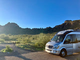 Silver Mercedes Sprinter RV rental parked at a desert campsite, surrounded by mountains & scrub under a clear blue sky.