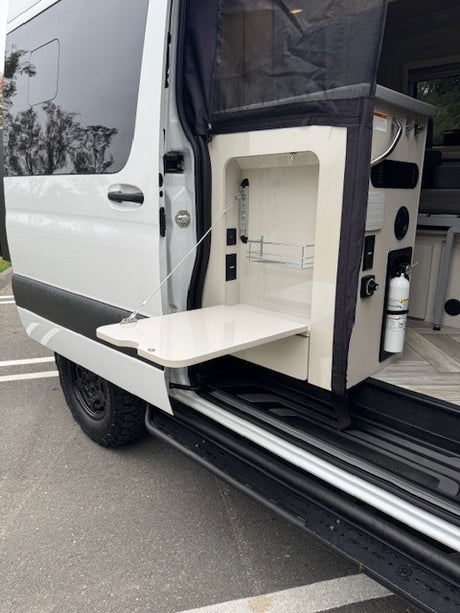 RV rental: Open side view of a camper van's exterior kitchen, featuring a pull-out table, storage, and fire extinguisher.