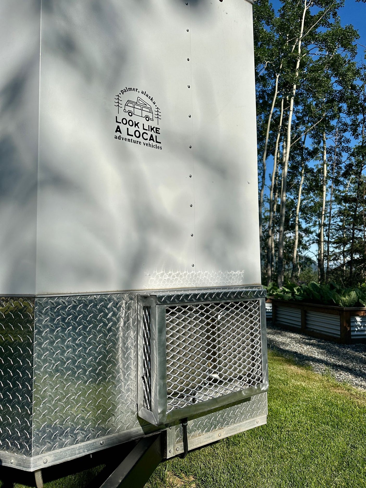 White RV rental trailer from "Look Like a Local" (Palmer, AK) with diamond plate trim, set in sunny Alaska nature.
