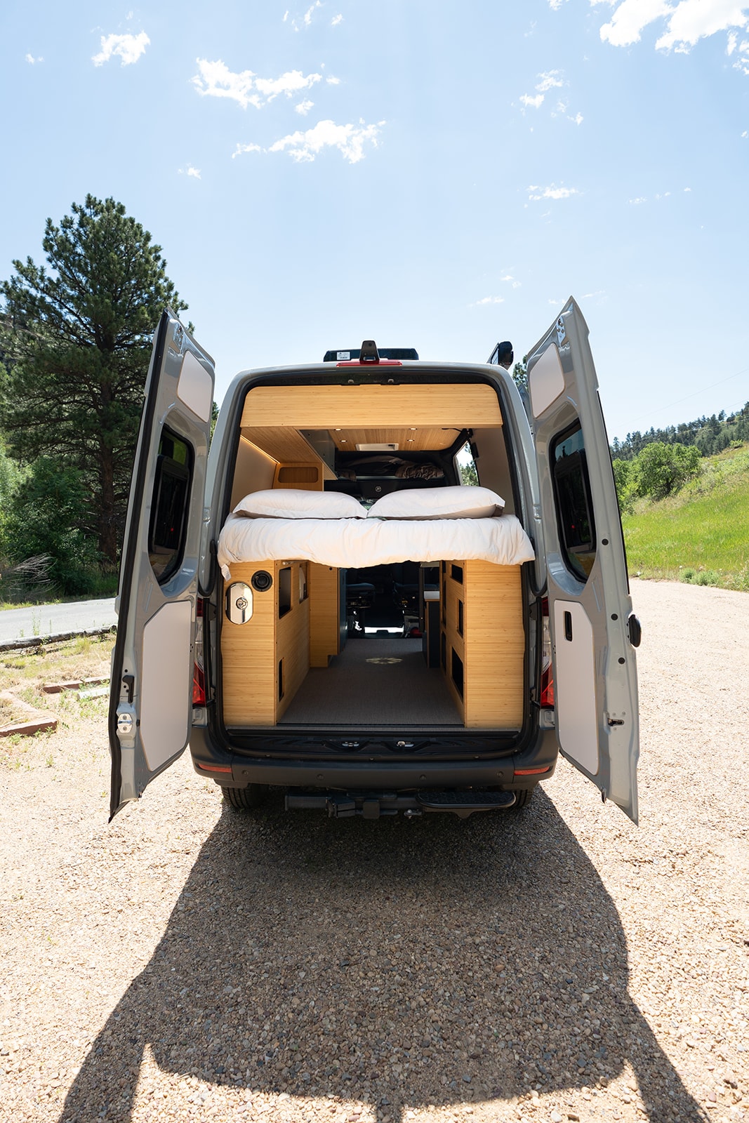 Custom RV rental camper van with open rear doors showing a cozy bed & bamboo interior, parked on a gravel road. — photo 18