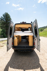 Custom RV rental camper van with open rear doors showing a cozy bed & bamboo interior, parked on a gravel road.