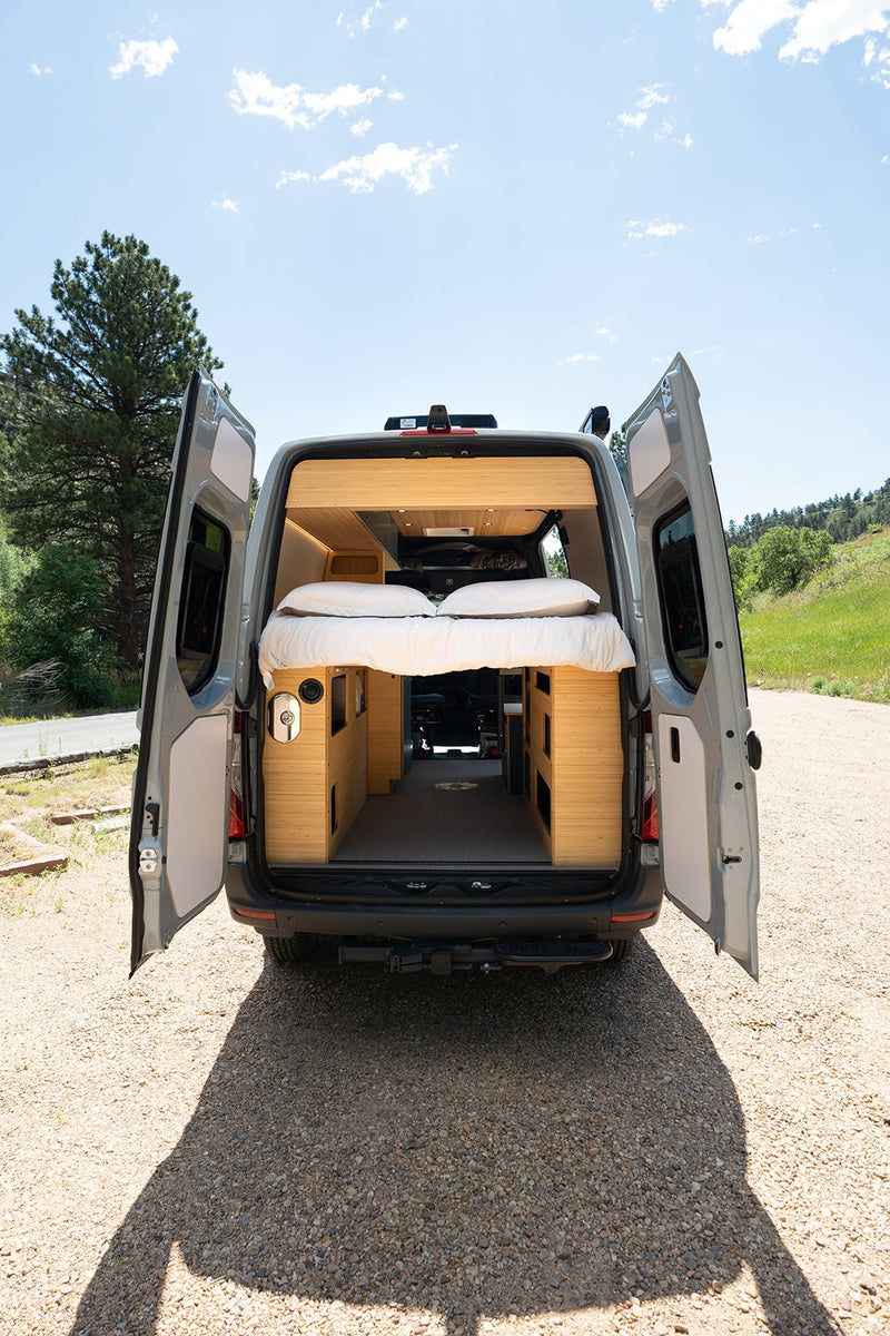 Custom RV rental camper van with open rear doors showing a cozy bed & bamboo interior, parked on a gravel road. — photo 18