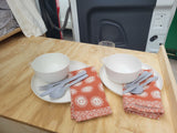 RV rental dining setup: two full place settings, with bowls, plates, cutlery, and sun-patterned napkins on a wooden table.