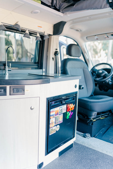 Inside an RV rental: a compact kitchen with sink, induction cooktop, and fridge adorned with travel magnets.