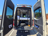 Open rear doors of an RV rental show a functional interior: elevated bed, under-bed storage with a spare tire, and door nets.