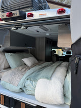Inside a comfy RV rental: a neat bed with soft bedding, overhead storage, and AC, viewed from the open rear door.